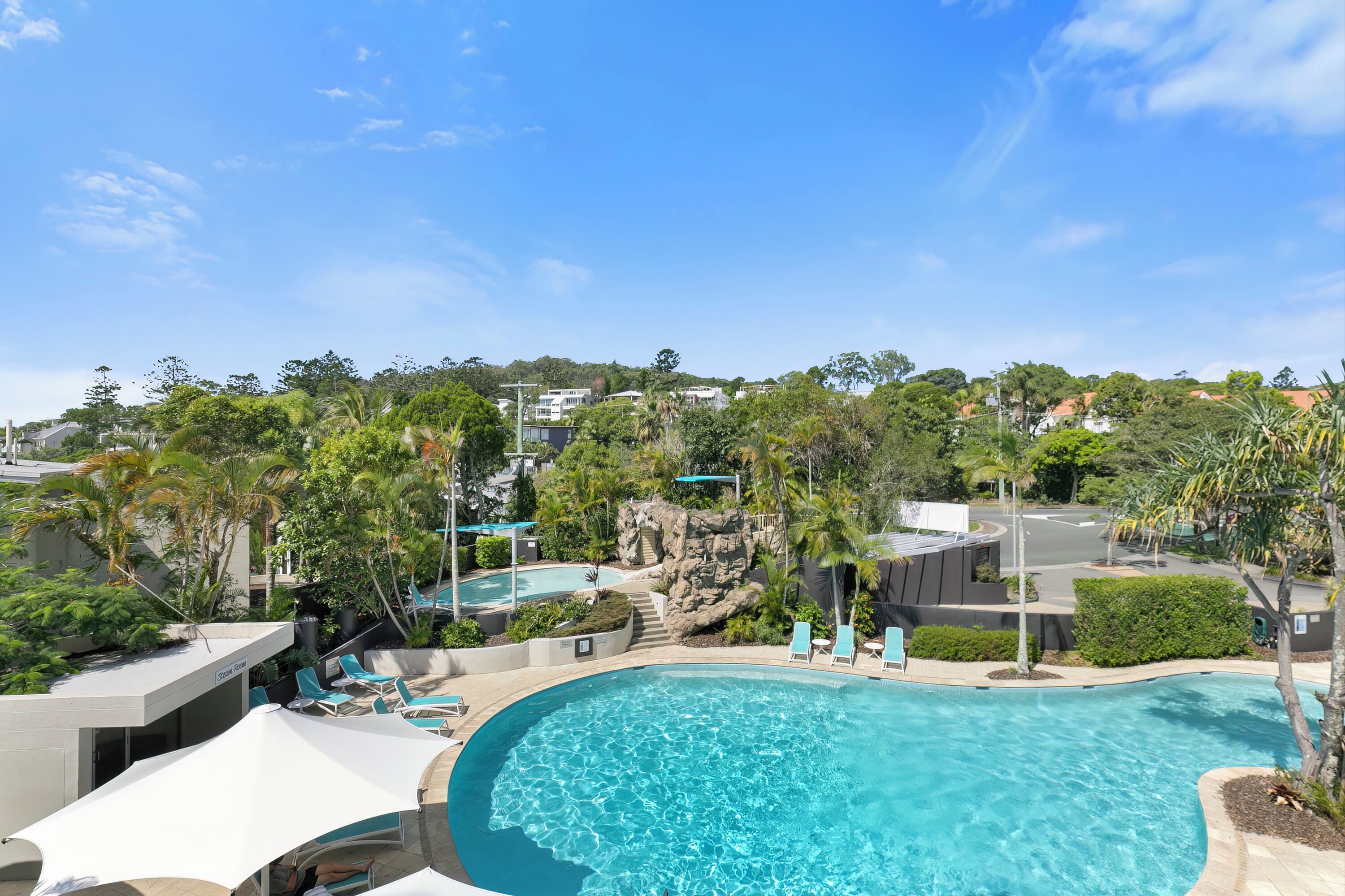 A swimming pool with lounge chairs and umbrellas, surrounded by greenery and buildings.
