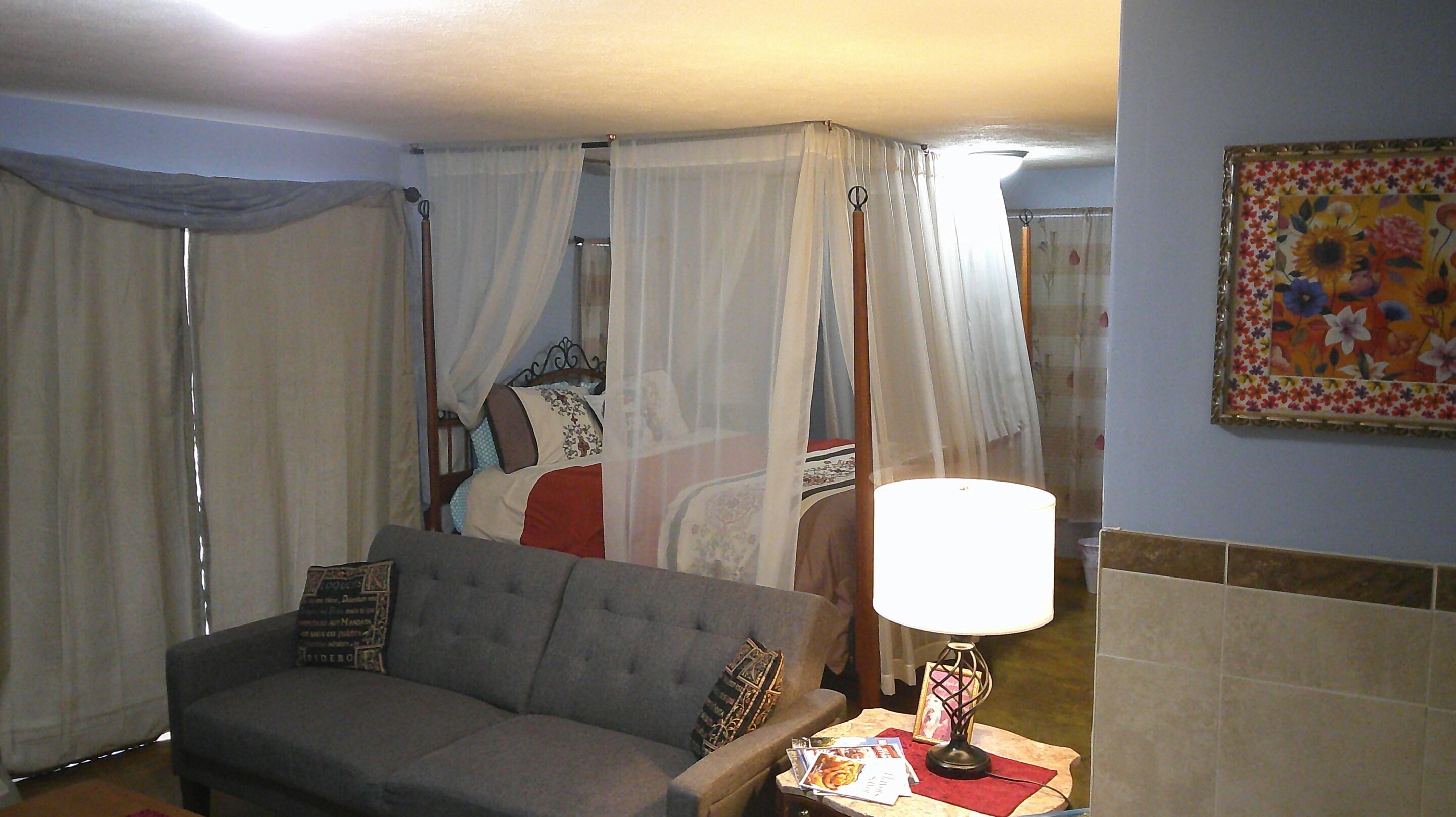 A hotel room with a grey sofa, a bedside table with a lamp, and a bed with a canopy.