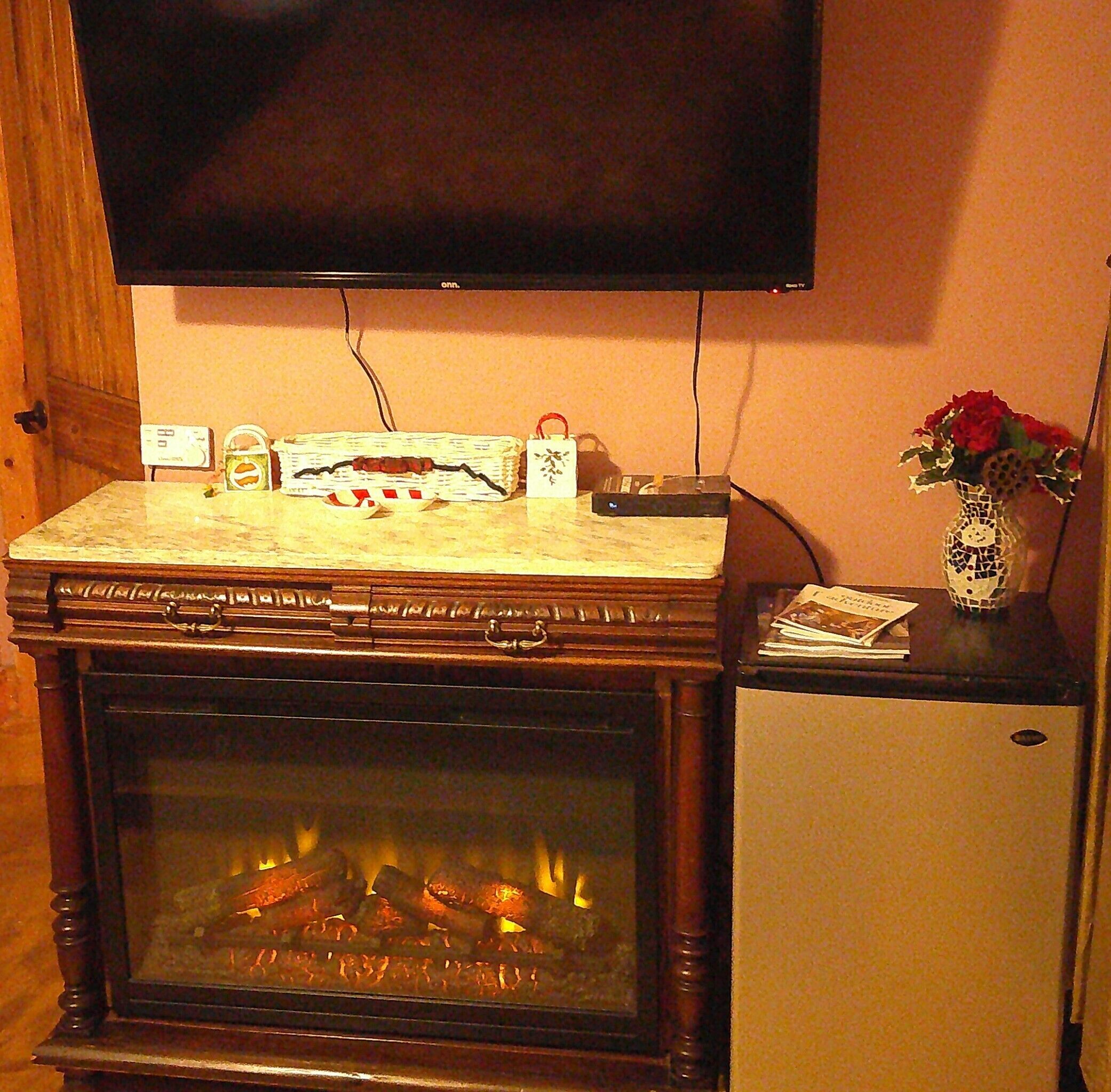Una chimenea con un televisor encima, una mesita con un jarrón de flores y un reloj en la pared.
