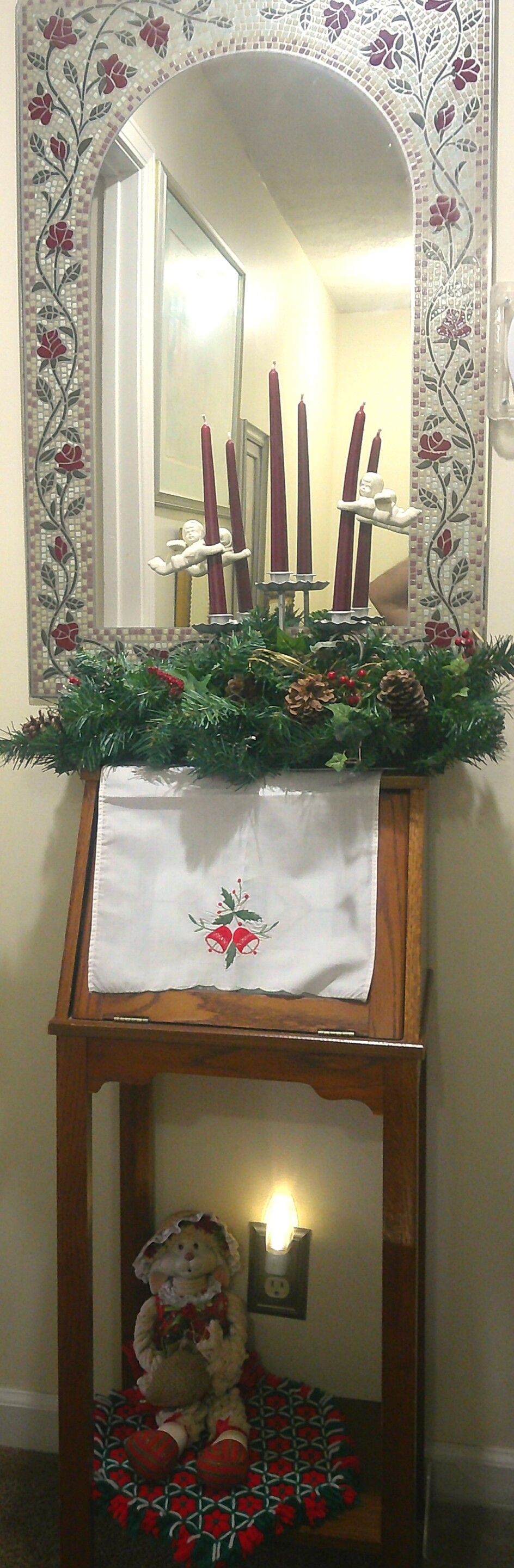 A wooden console table with a white cloth and holly decoration, a mirror with floral mosaic, and a teddy bear on a red and green patterned cushion.
