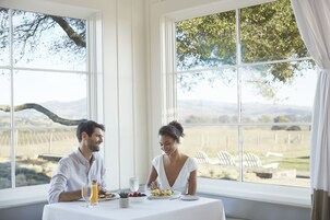 Una mesa de comedor con comida servida, un vaso de jugo de naranja y vista a un viñedo a través de amplias ventanas.