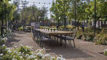 An outdoor dining area with a long wooden table and chairs, surrounded by lush greenery and flowering plants.