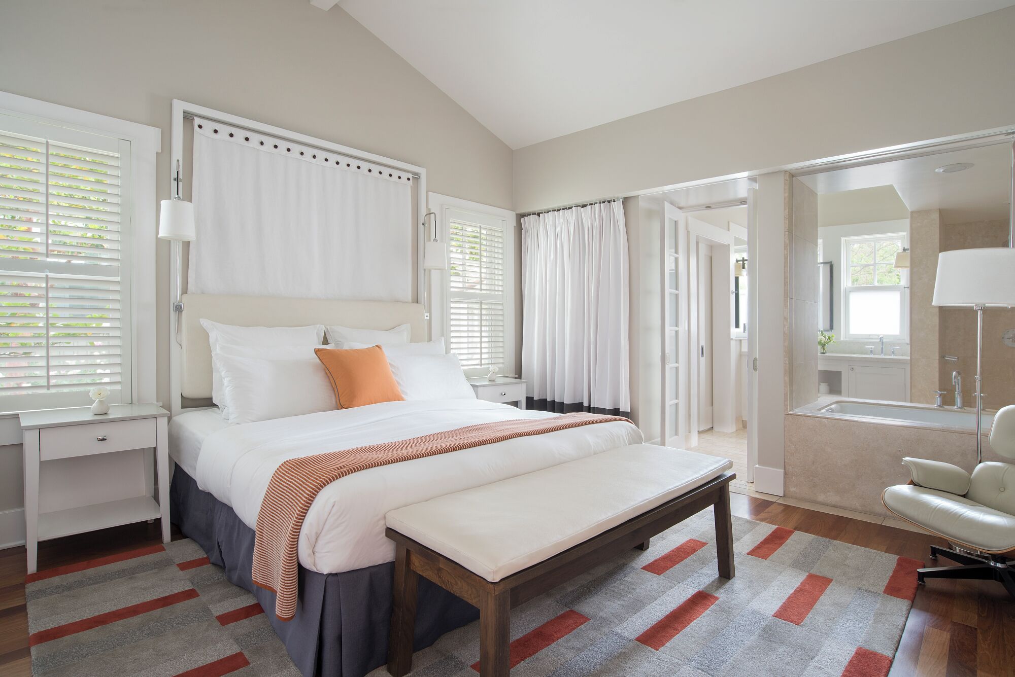A bedroom with a large bed, a bench, a chair, and a view of the bathroom.
