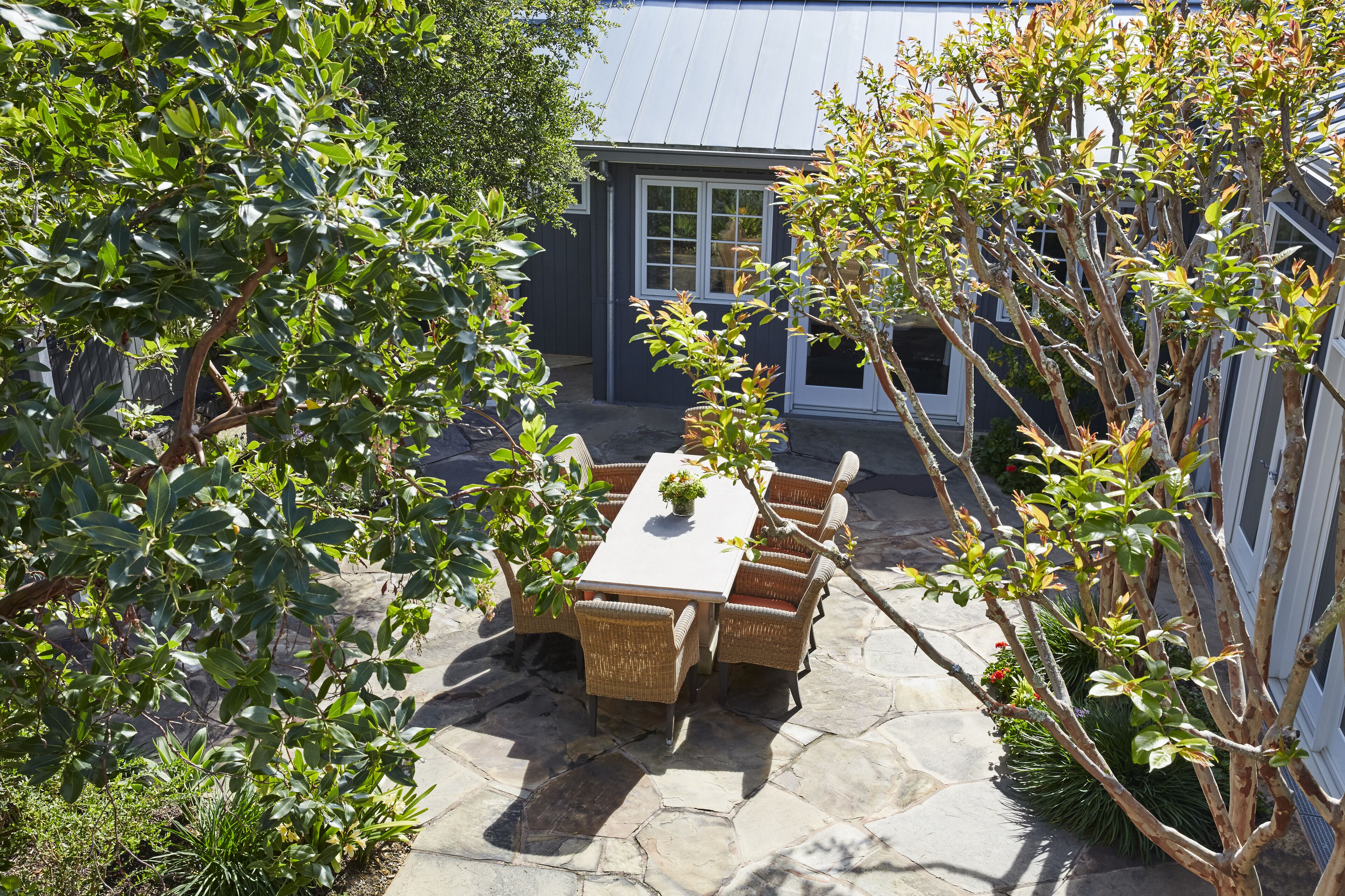 A patio with a table and chairs, surrounded by greenery and a house in the background.