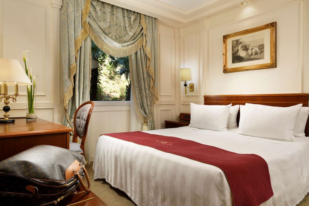 Quarto de hotel com uma cama grande, uma escrivaninha com abajur, uma cadeira e uma janela com cortinas.