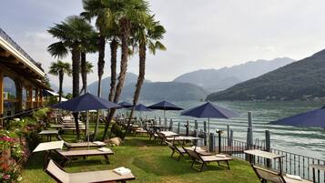 A lakeside patio with tables and chairs, umbrellas, and palm trees.