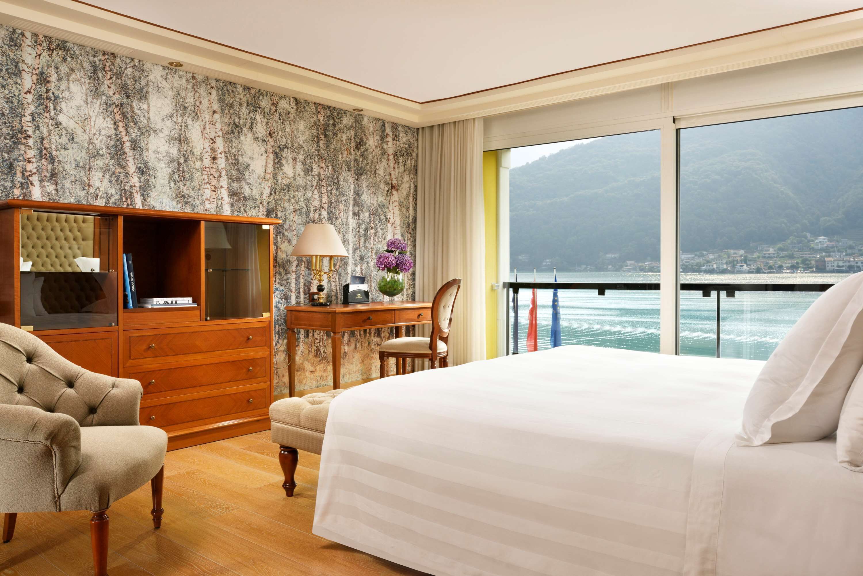 Ein Hotelzimmer mit einem großen Bett, einem hölzernen Schminktisch, einem Sessel, einem Schreibtisch mit Lampe und Blick auf ein Gewässer und Berge.