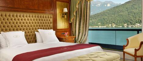 A luxurious hotel room with a large bed, a wooden headboard, a view of a lake and mountains, and a balcony with a chair.