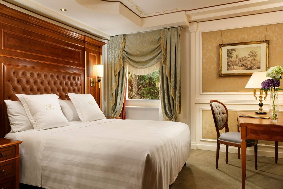 Quarto de hotel com uma cama grande, cabeceira de madeira, escrivaninha com cadeira e janela com cortinas.