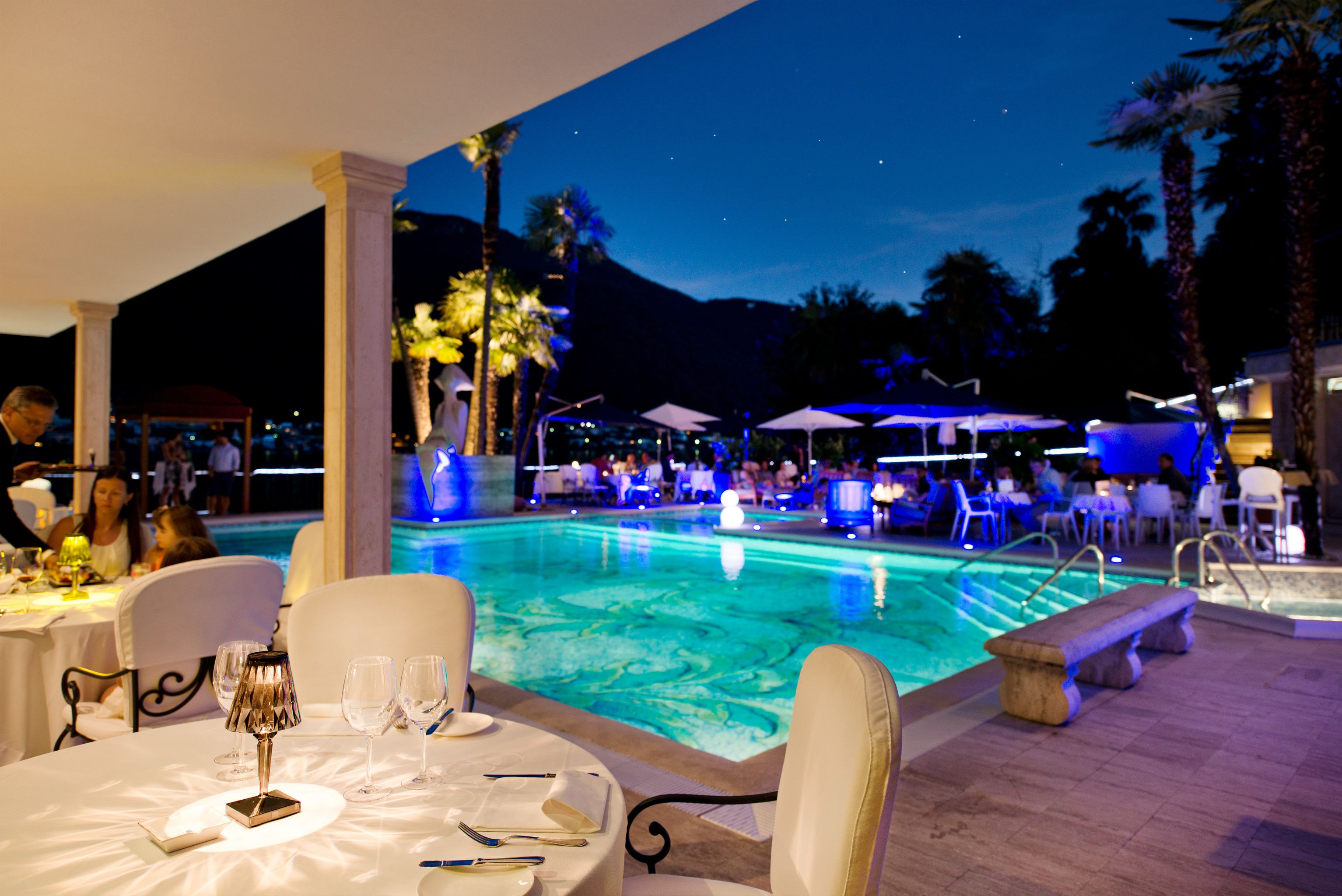 A poolside dining area with tables set for guests, a pool with clear water, and palm trees in the background.