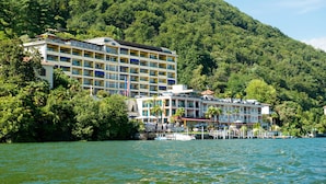 Ein mehrstöckiges Hotel mit Balkonen, gelegen an einem See mit einer Anlegestelle und Booten, umgeben von üppigen grünen Hügeln.