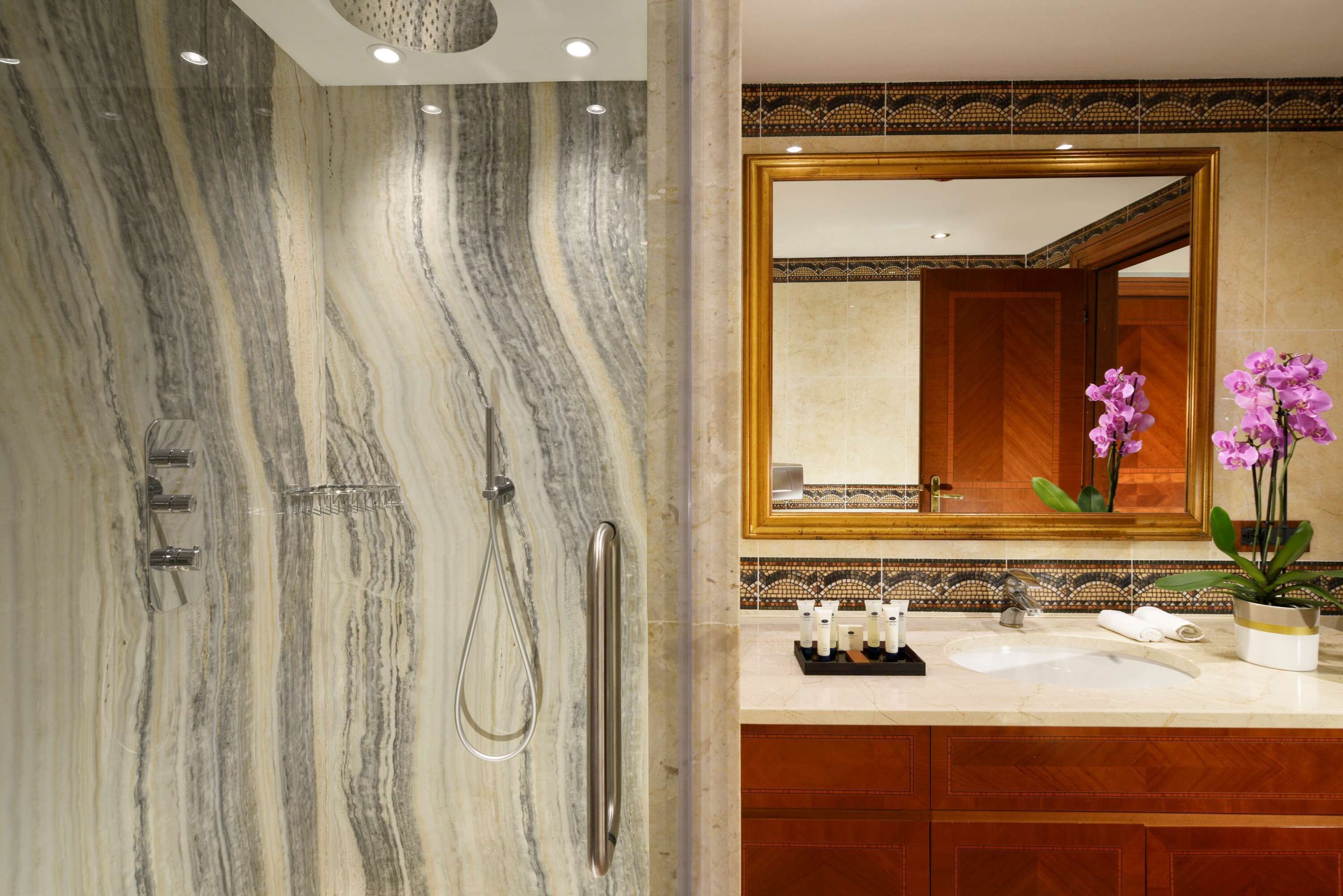 Un baño con pared de ducha de mármol, un espejo grande y un lavamanos con artículos de aseo.