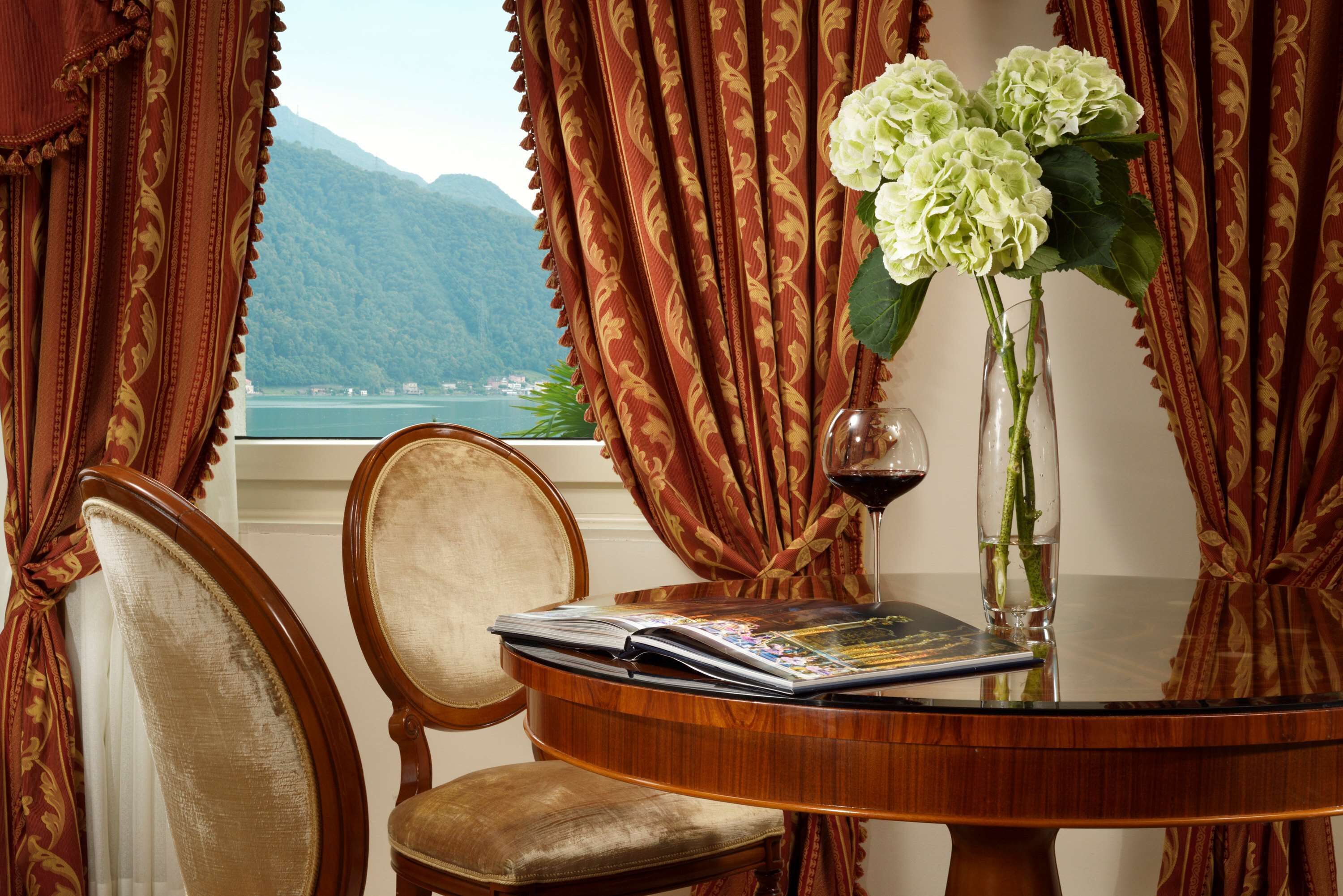 A room with a view of mountains and a lake, a table with a magazine, a vase with flowers, and a wine glass.