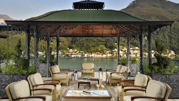 Un espace extérieur aménagé, abrité et doté de chaises en osier et d’une table dressée pour un repas, offrant une vue sur un lac et des montagnes.