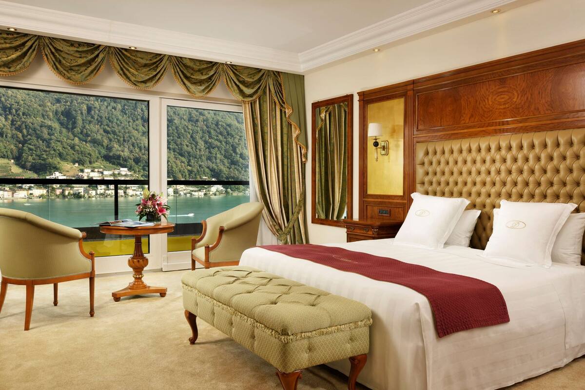 Quarto de hotel com uma cama grande, um espaço de estar com uma poltrona e uma mesa, e vista para uma área arborizada por grandes janelas.