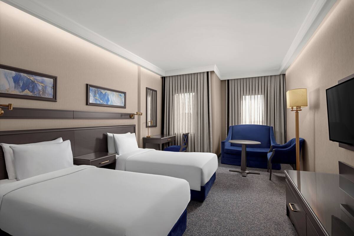 A hotel room with two beds, a desk, a chair, a TV, and a window with curtains.