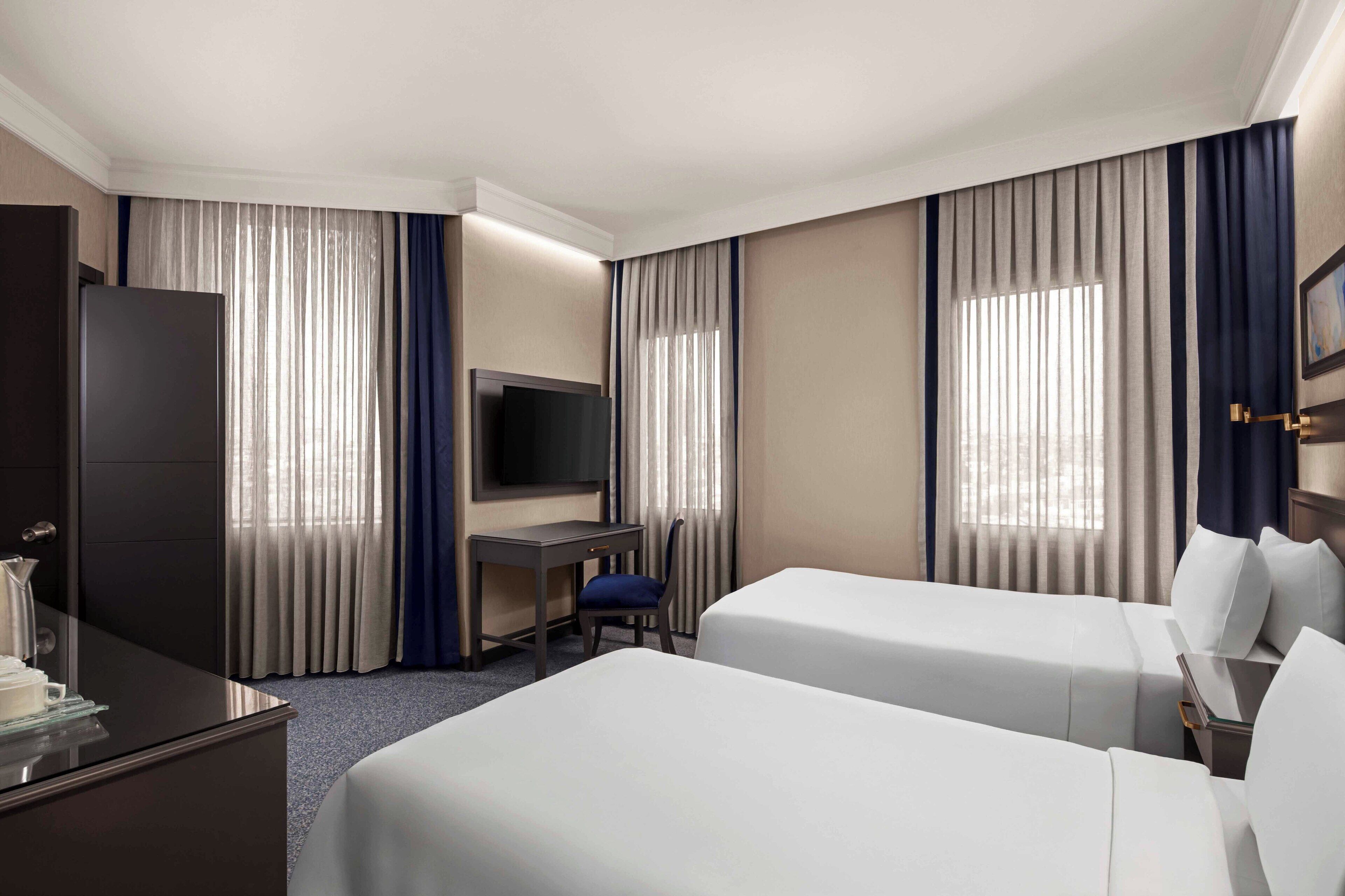 A hotel room with two beds, a desk, a TV, and curtains.