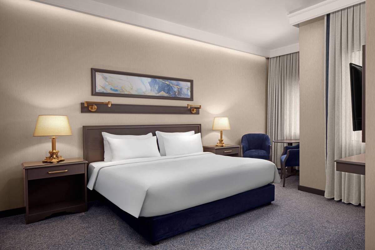 A hotel room with a large bed, two bedside tables with lamps, a chair, a small table, and a TV.