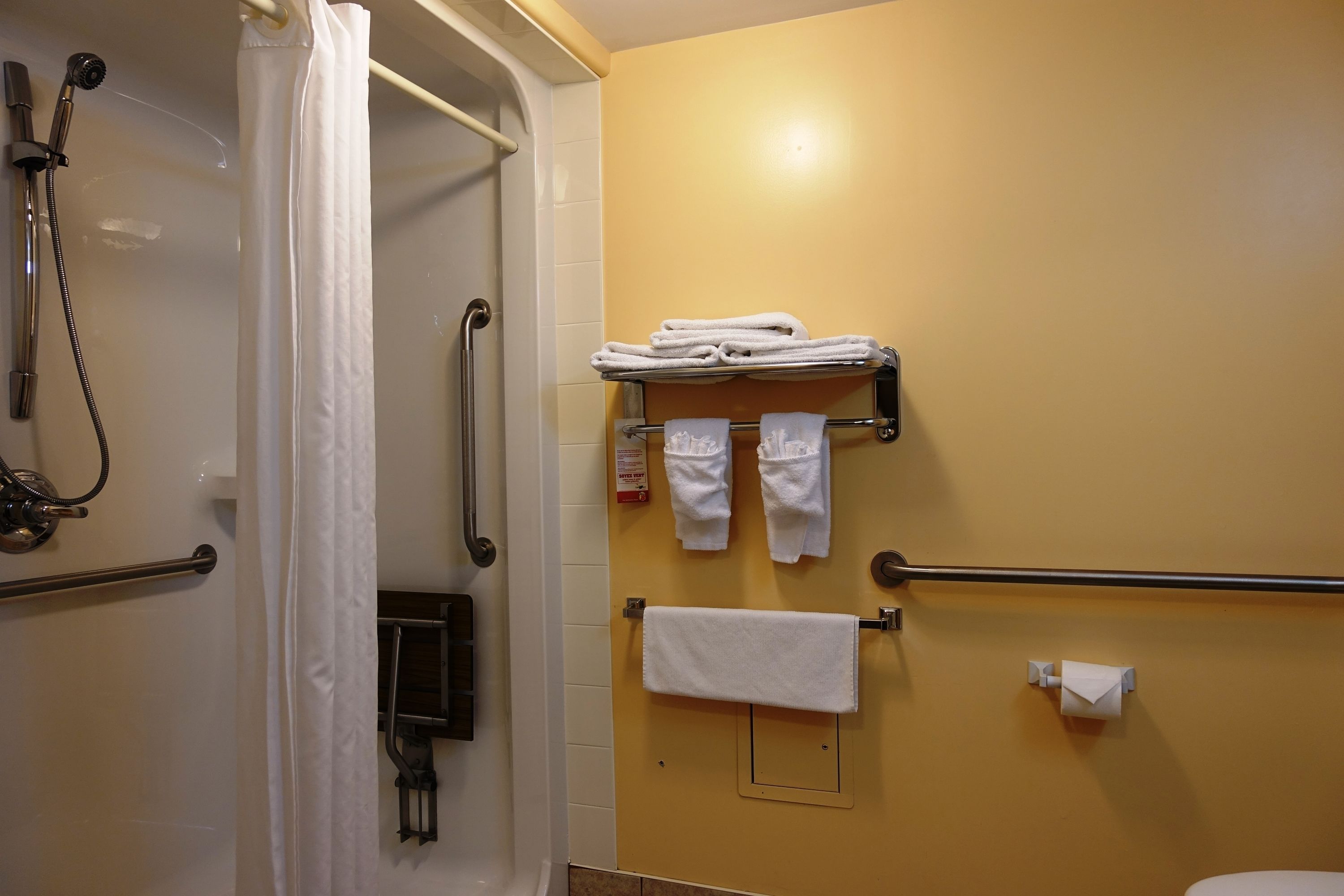 Une salle de bain équipée d’une douche, de barres d’appui, d’un porte-serviettes avec des serviettes pliées, d’un support à papier toilette et d’un autre porte-serviettes fixé au mur.
