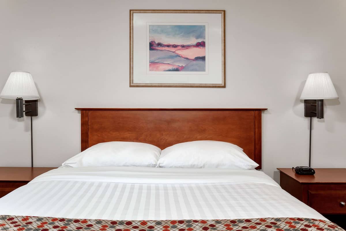 A neatly made bed with white linens, a wooden headboard, and a framed painting above it.