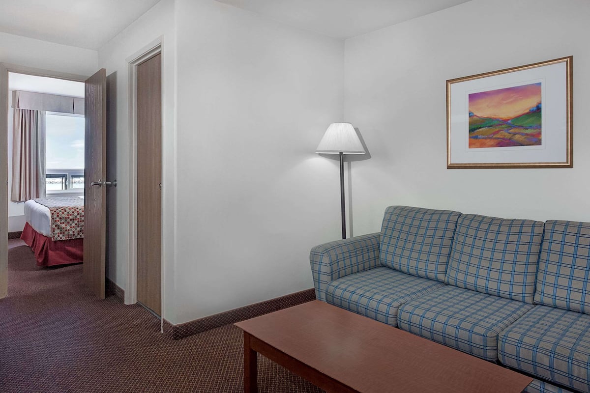 A hotel room with a checkered sofa, a wooden table, a lamp, a painting on the wall, and a glimpse of a balcony with a bed.
