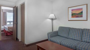A hotel room with a checkered sofa, a wooden table, a lamp, a painting on the wall, and a glimpse of a balcony with a bed.