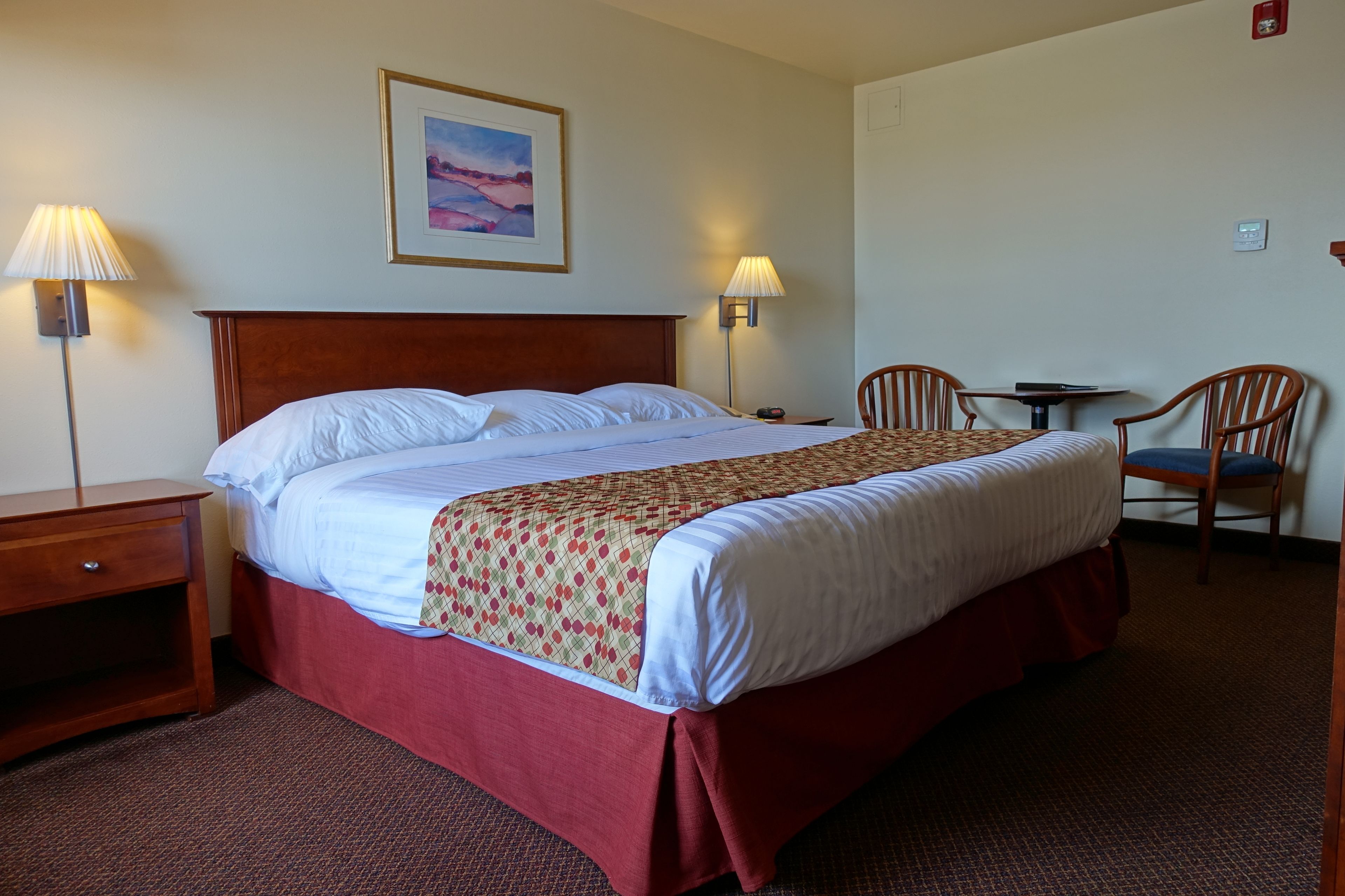 A hotel room with a large bed, two bedside lamps, a small table with chairs, and a framed picture on the wall.