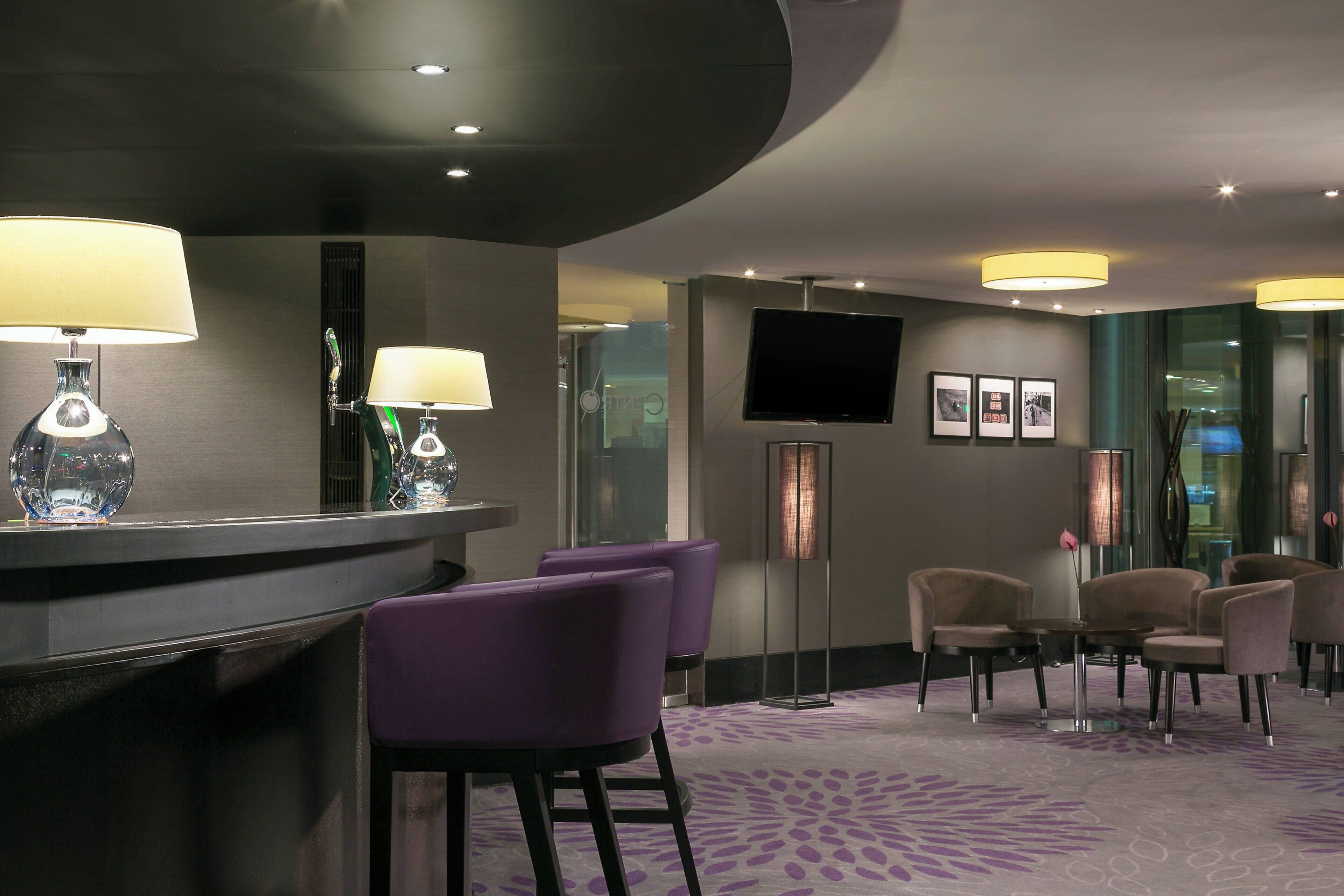 Um hall de hotel com balcão de recepção, assentos em tons de roxo e bege, uma TV de tela plana e quadros nas paredes.