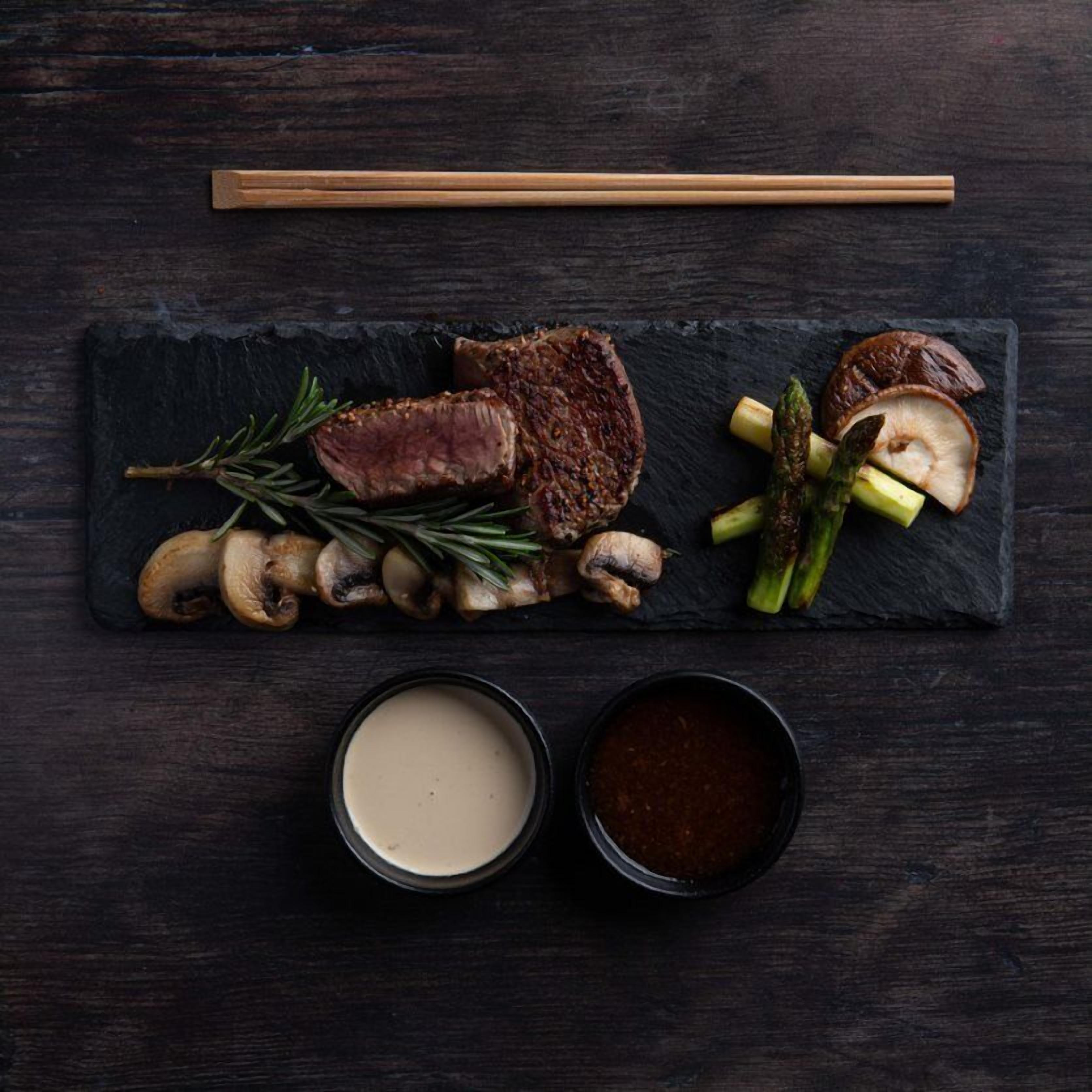 A dark wooden table with a black slate serving board holding sliced steak, mushrooms, asparagus, and rosemary. Two small bowls of sauce are also visible.