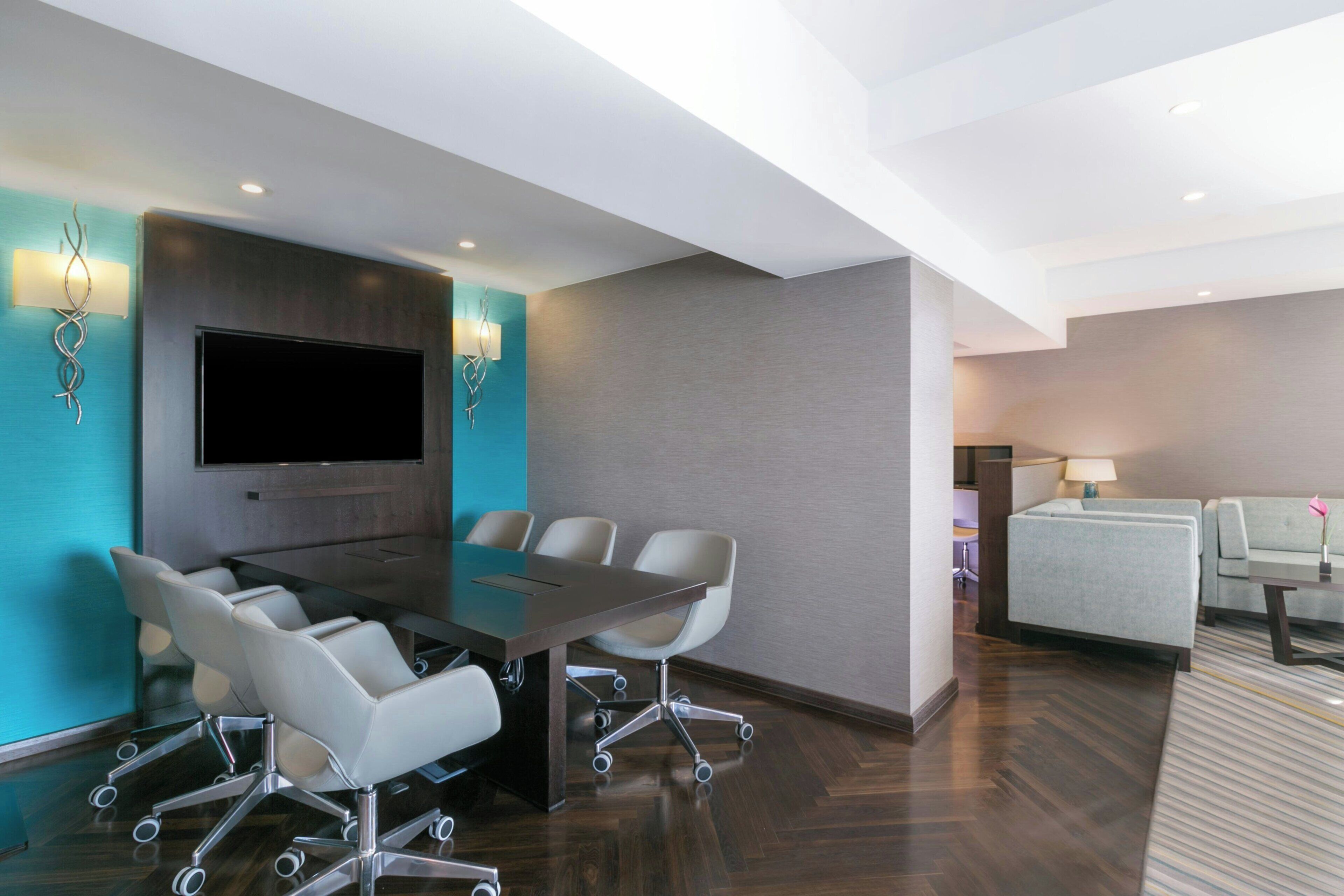 A modern conference room with a large table, white chairs, a flat-screen TV, and a sofa in the adjacent living area.