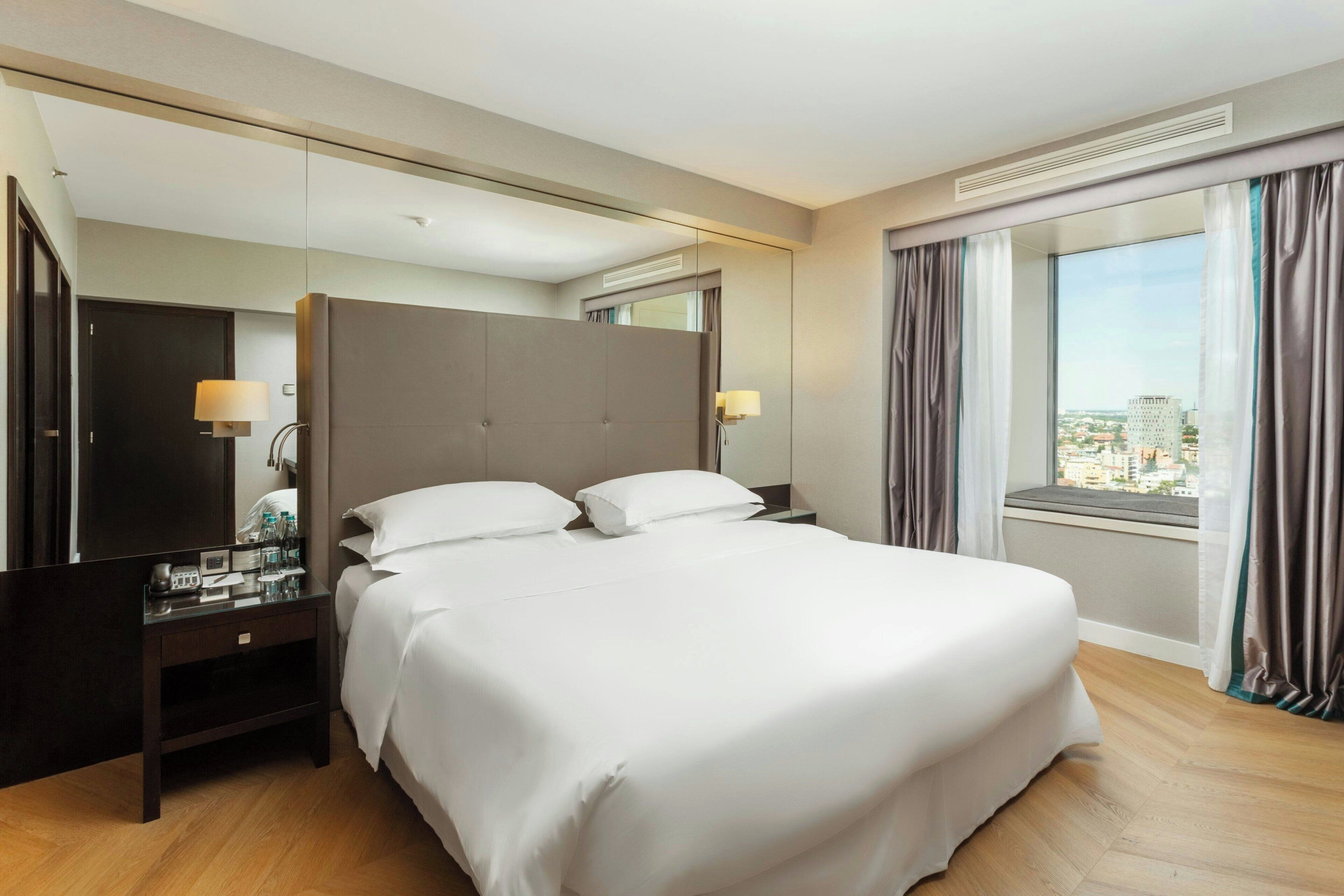 A hotel room with a large bed, a bedside table with a lamp, a mirror, and a window with curtains.