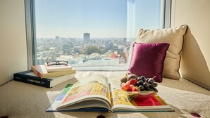 Een raamzitplaats met uitzicht op een stadsgezicht, een boek getiteld 'The World Travel Atlas', een paar brillen en een schaal fruit.