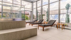 Een glazen loungeruimte met zachte zitplaatsen en een tafeltje, met uitzicht op de omliggende gebouwen.