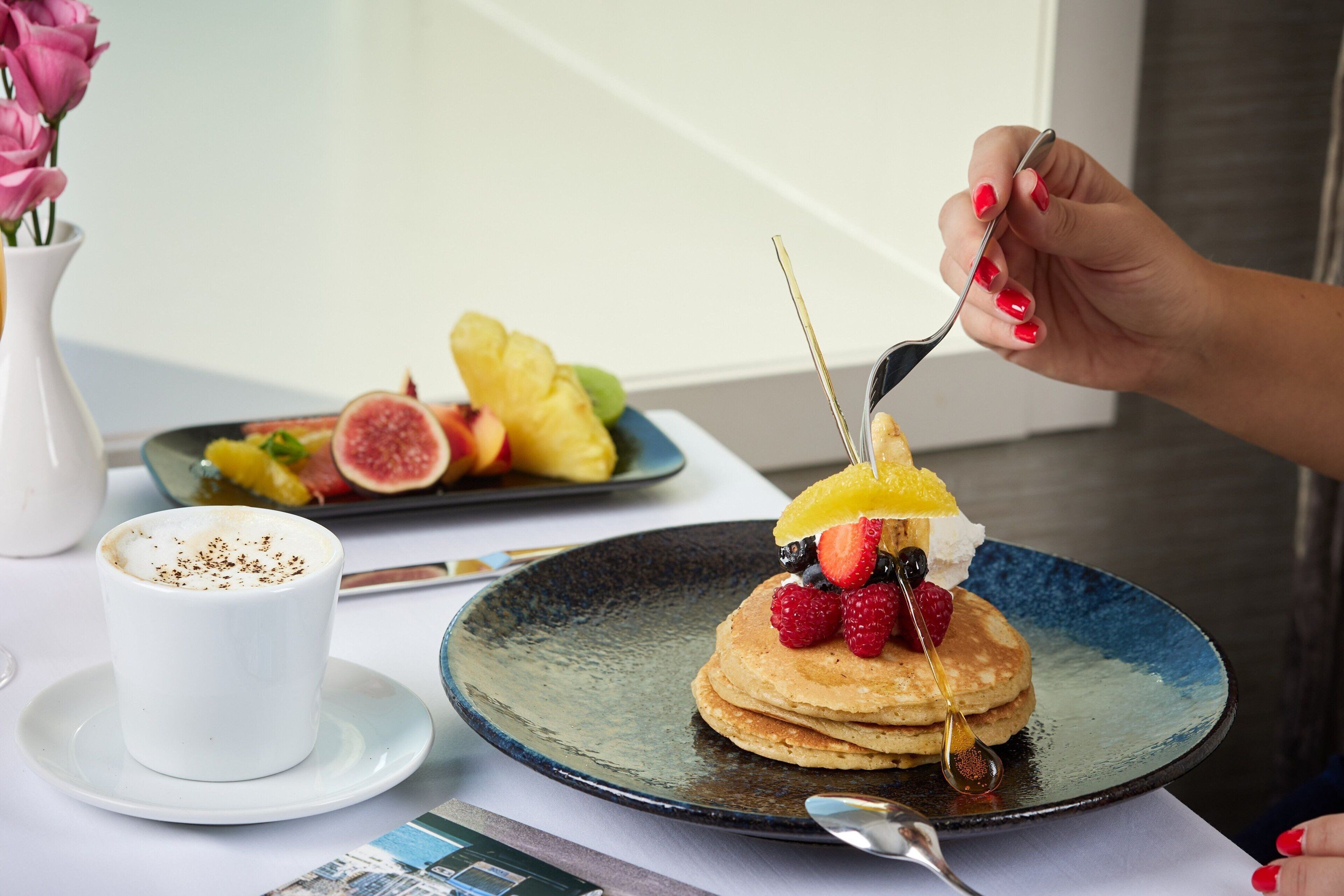 Ein Frühstückstisch mit Pfannkuchen, Obst und Sirup, eine Tasse Kaffee und einem Teller Croissants.