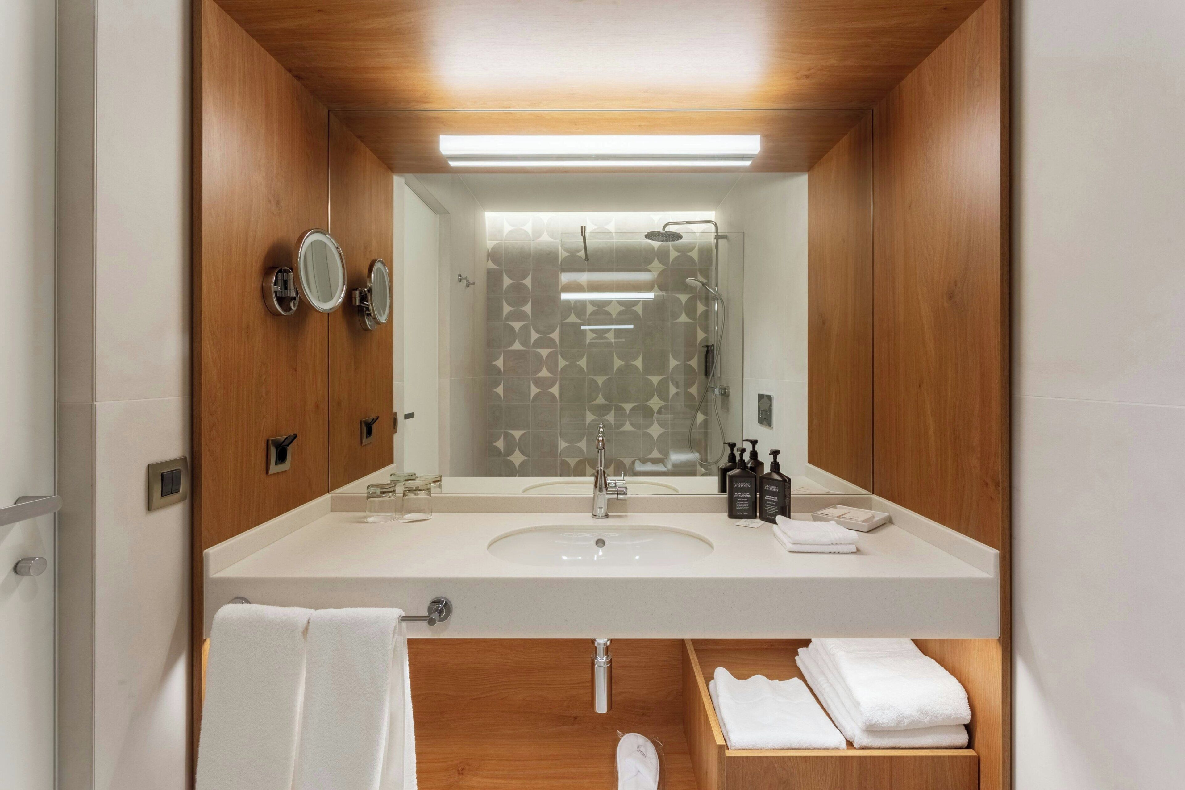 A modern bathroom with a wooden vanity, a large mirror, a shower with a geometric tile pattern, and a white sink.