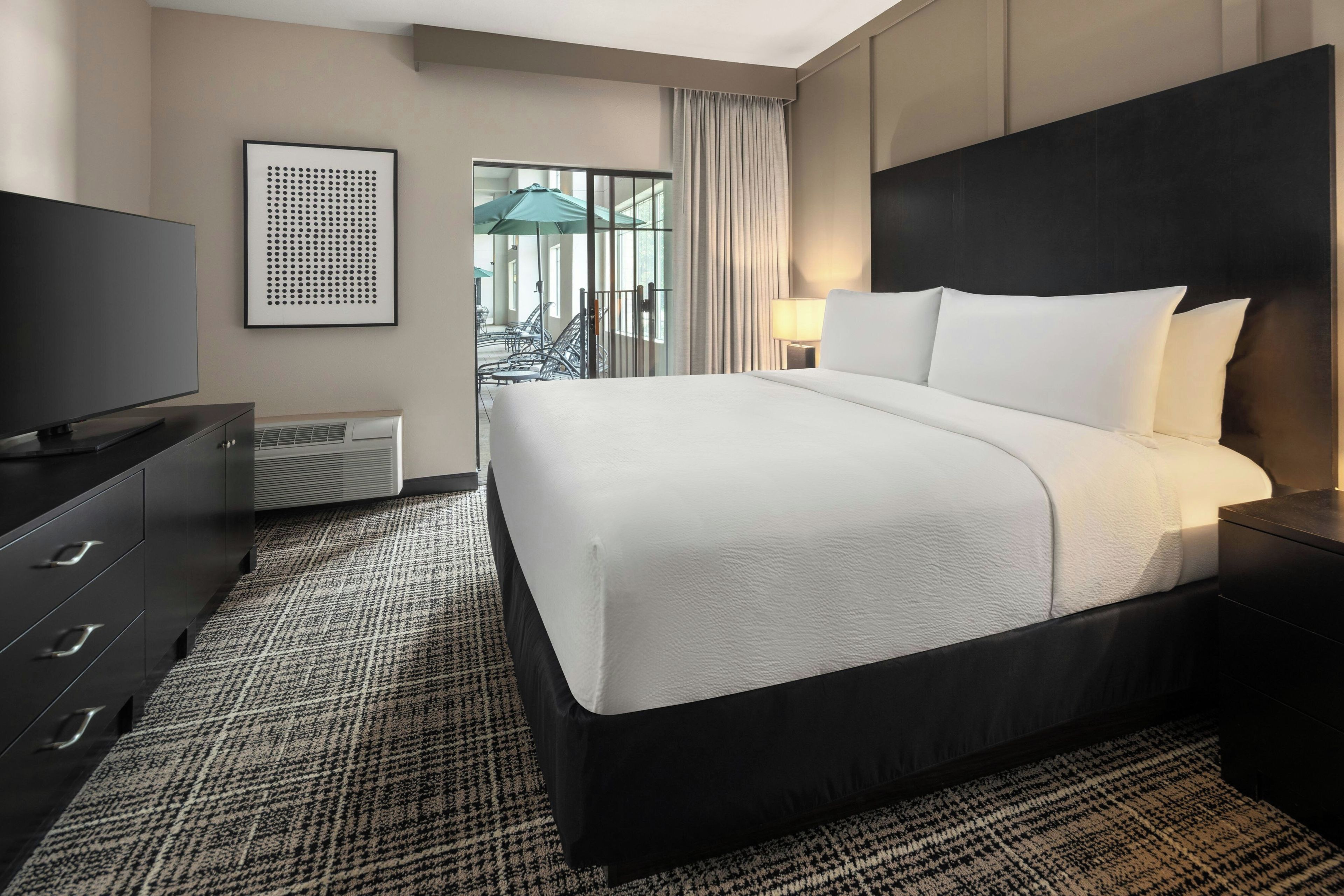 A hotel room with a large bed, a television, a dresser, and a view of the outdoor area.