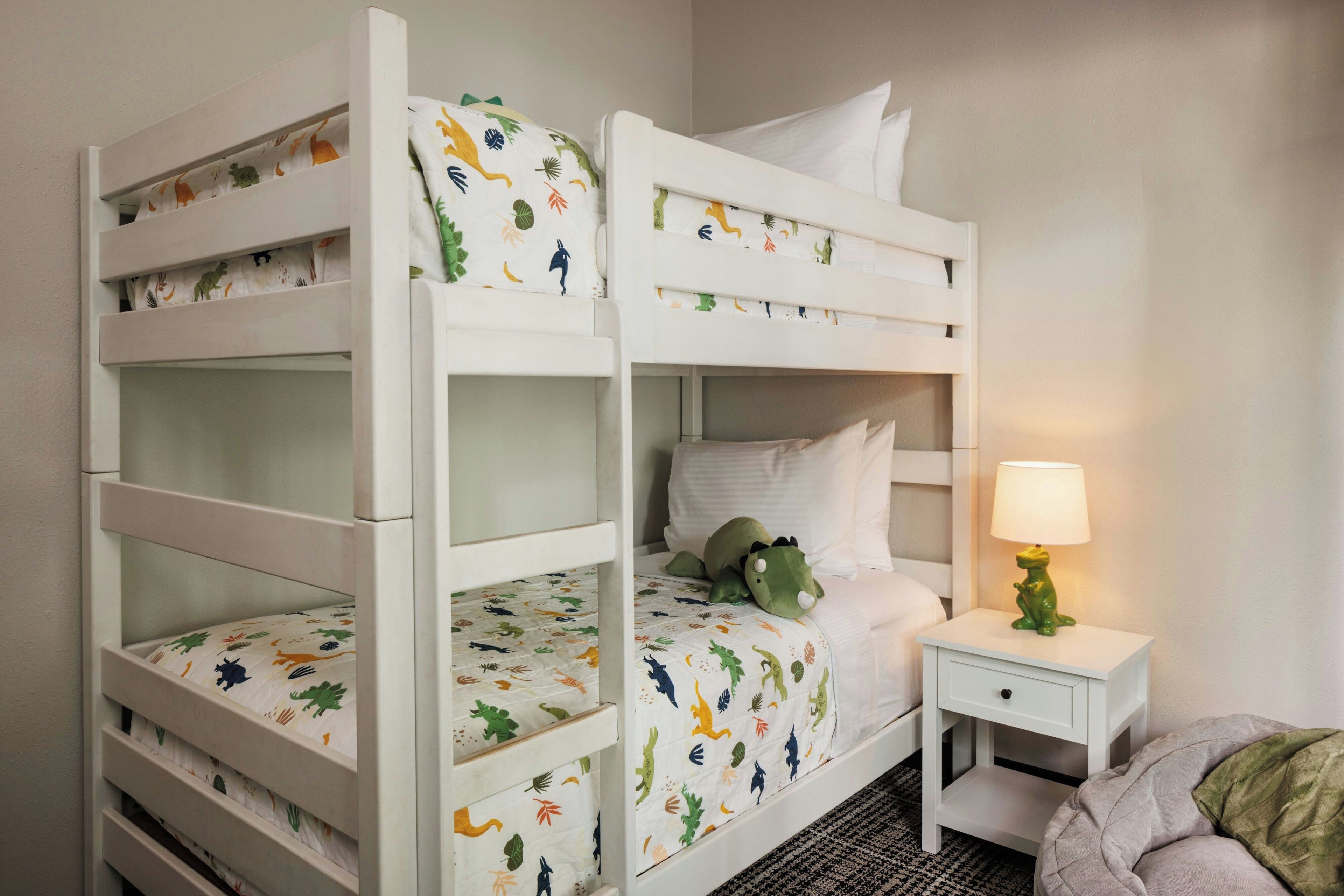 A bunk bed with a dinosaur-themed bedspread, a bedside lamp, and a small bedside table.