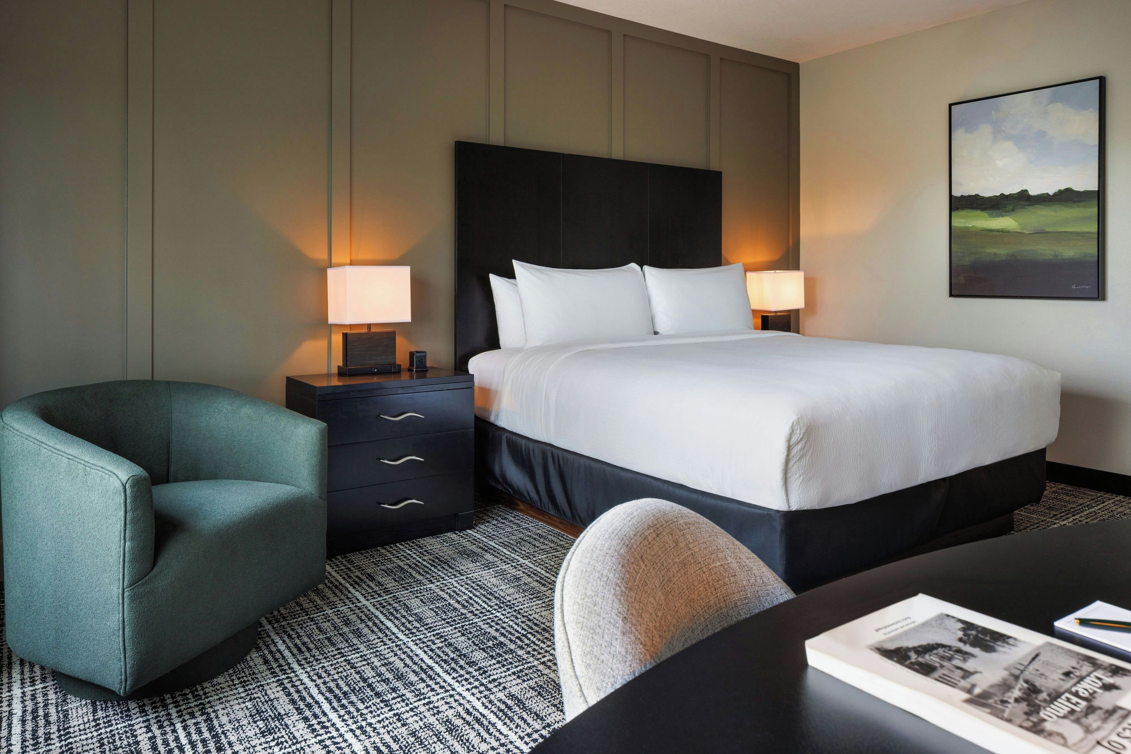 A hotel room with a bed, two bedside tables with lamps, a chair, a desk with a magazine, and a painting on the wall.