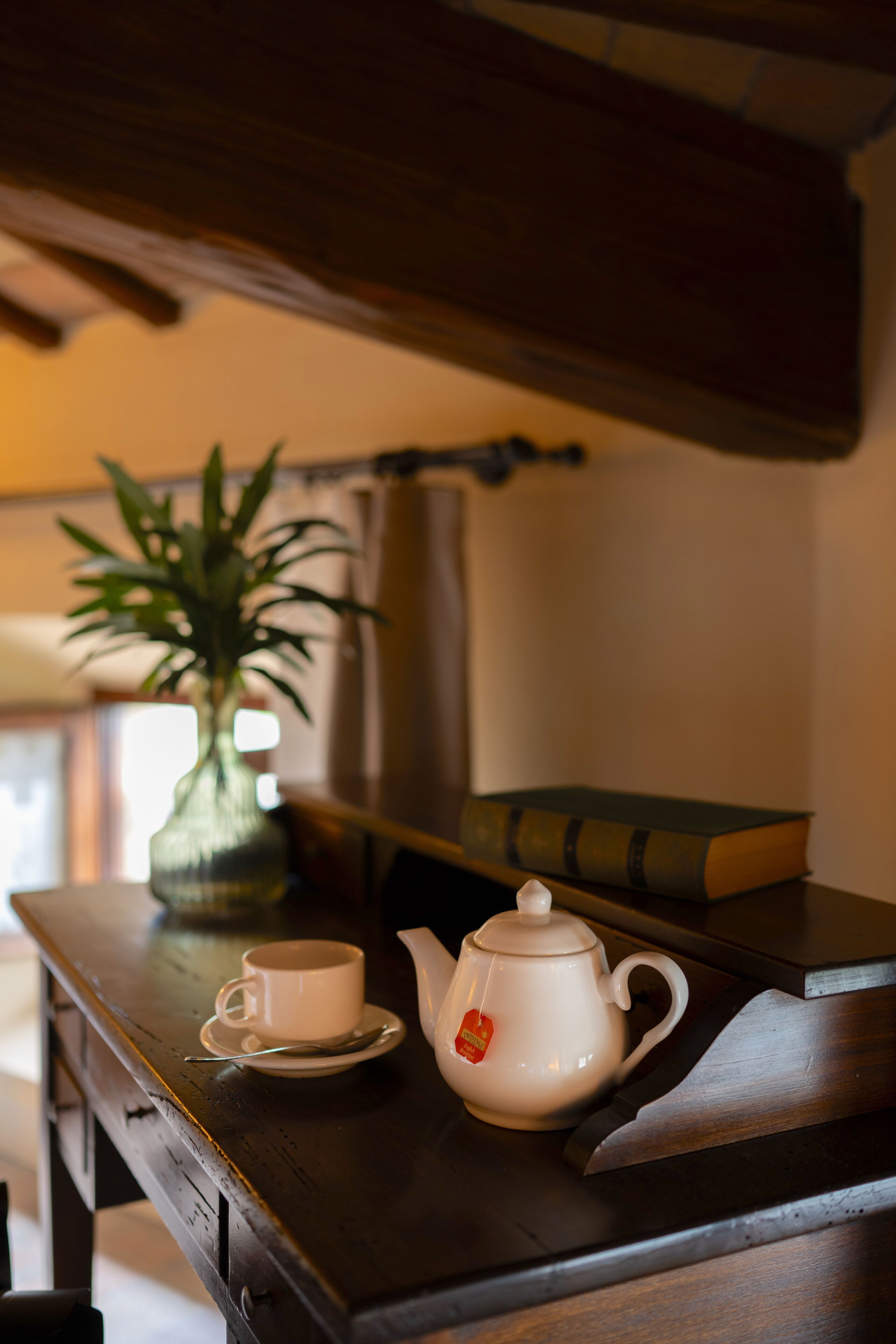 Ein Holztisch mit einem weißen Teekessel, einer Tasse und einem Untertasse. Auf dem Tisch liegen ein Buch und ein Topfpflanze.