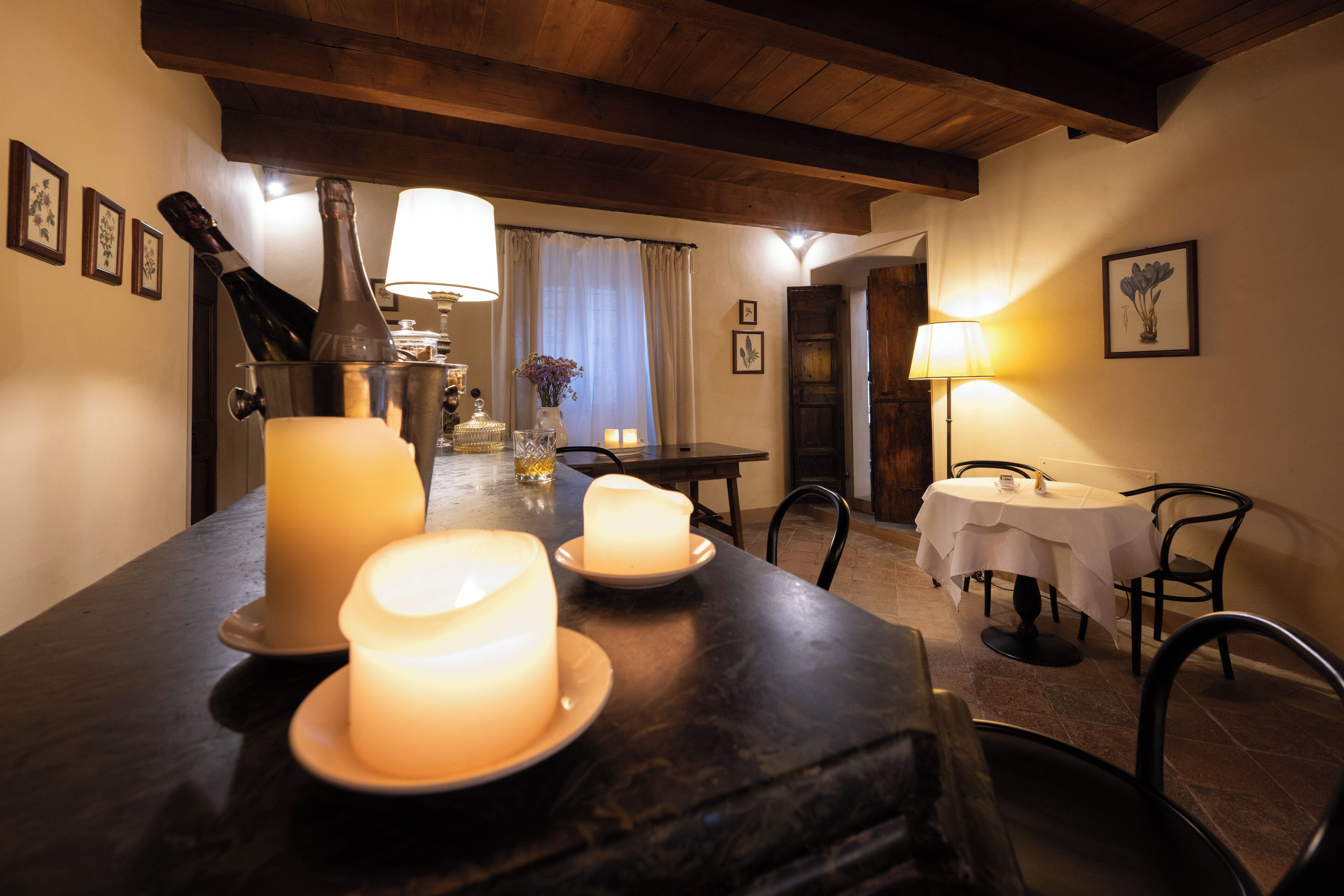 Un coin repas avec une longue table dressée pour deux personnes, des bougies, une bouteille de champagne et une lampe.