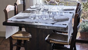 Tavola da pranzo imbandita per sei, con calici da vino, tovaglia bianca e vista sulle dolci colline attraverso una finestra.