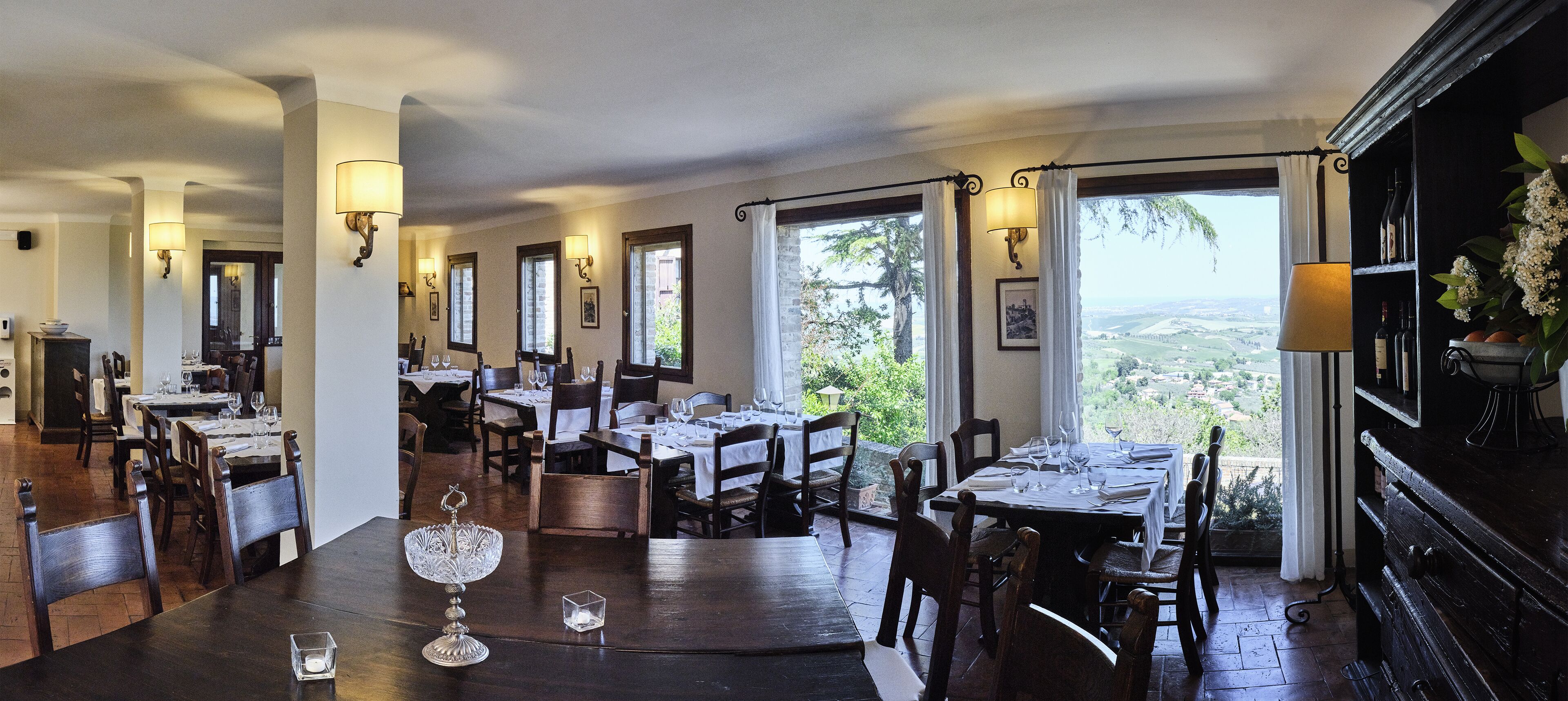 A dining area with multiple tables and chairs, large windows with curtains, and wall-mounted lamps.