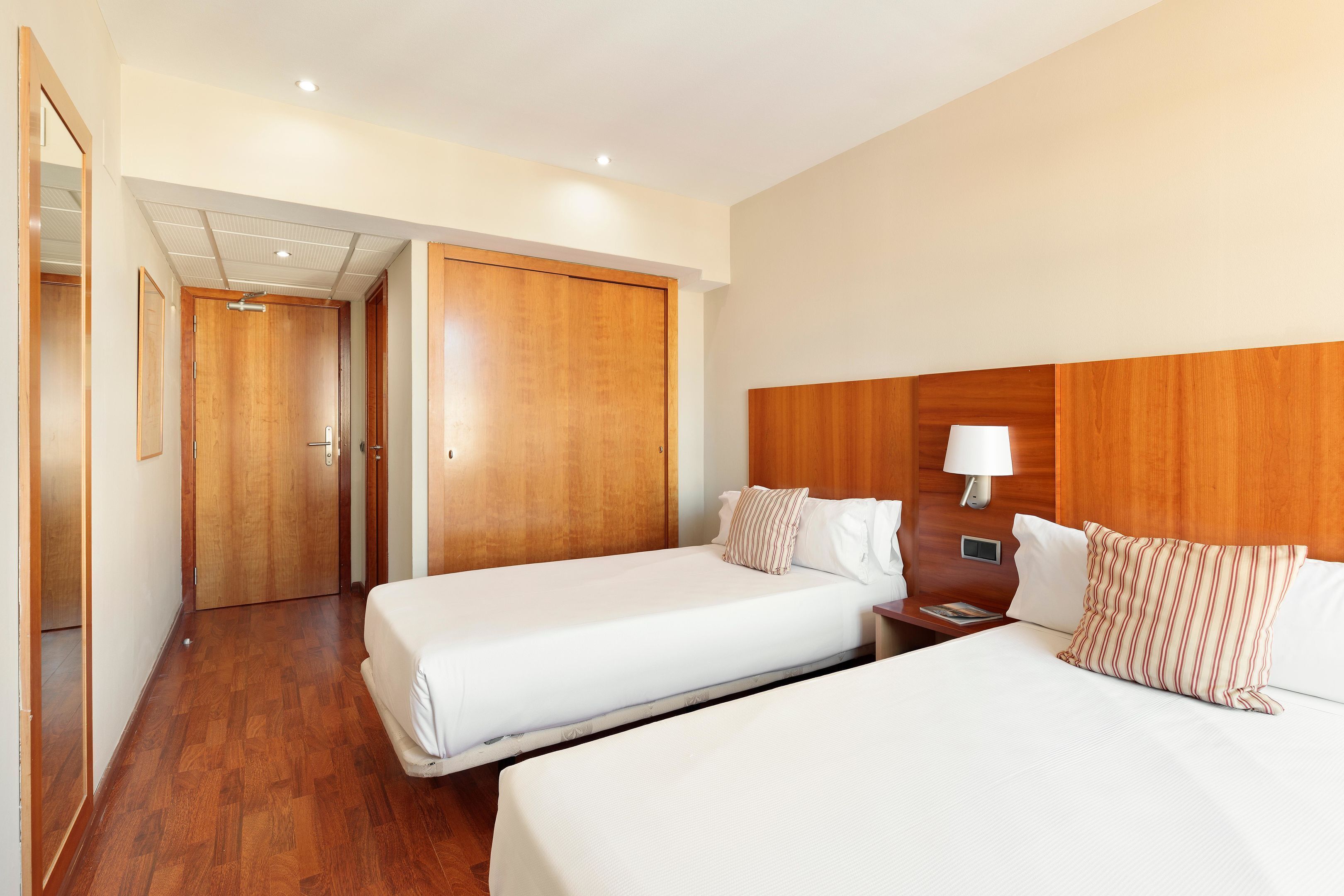 Habitación de hotel con dos camas, cabeceros de madera, una mesita de noche con lámpara y un armario de madera.