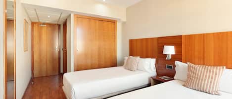 A hotel room with two beds, wooden headboards, a nightstand with a lamp, and a wooden wardrobe.