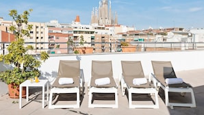 A rooftop lounge area with sun loungers and a potted plant, overlooking a cityscape with a prominent building in the background.