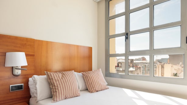 A hotel room with a wooden headboard, a bedside table with a lamp, two striped pillows, and a large window with a city view.