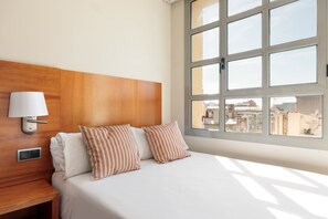 Habitación de hotel con cabecera de madera, mesita de noche con lámpara, dos almohadas a rayas y un ventanal con vistas a la ciudad.