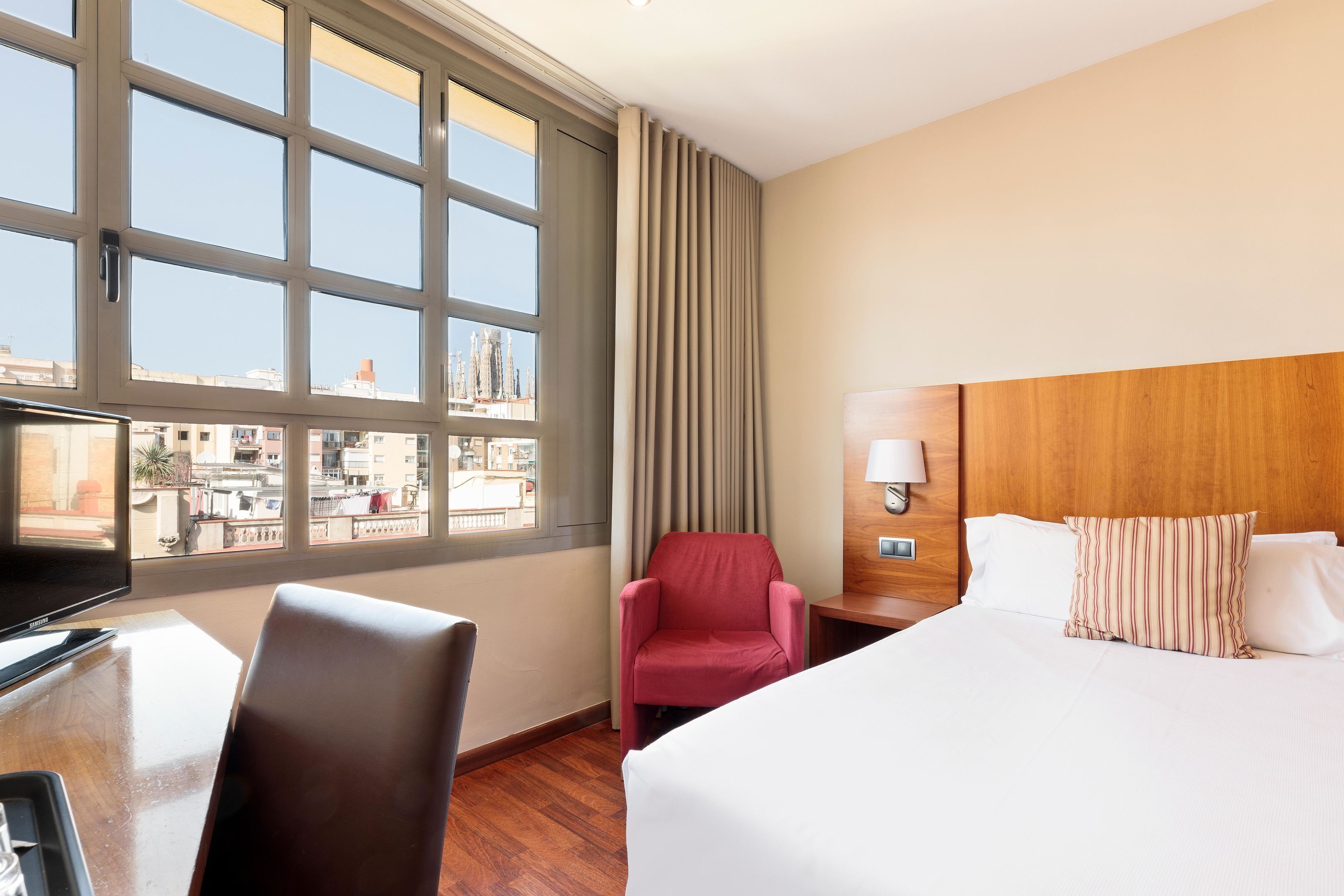 Habitación de hotel con cama, escritorio con computadora, silla, televisión y ventana con cortinas.