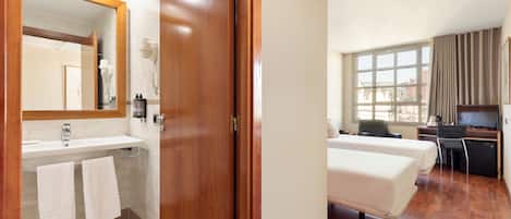 Una habitación de hotel con puerta de madera, un baño con lavabo y espejo, una cama, un escritorio con silla y una ventana con cortinas.