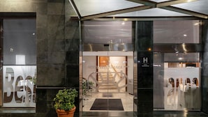 A hotel entrance with a modern design, featuring a glass door, a potted plant, and a visible reception area with a staircase.